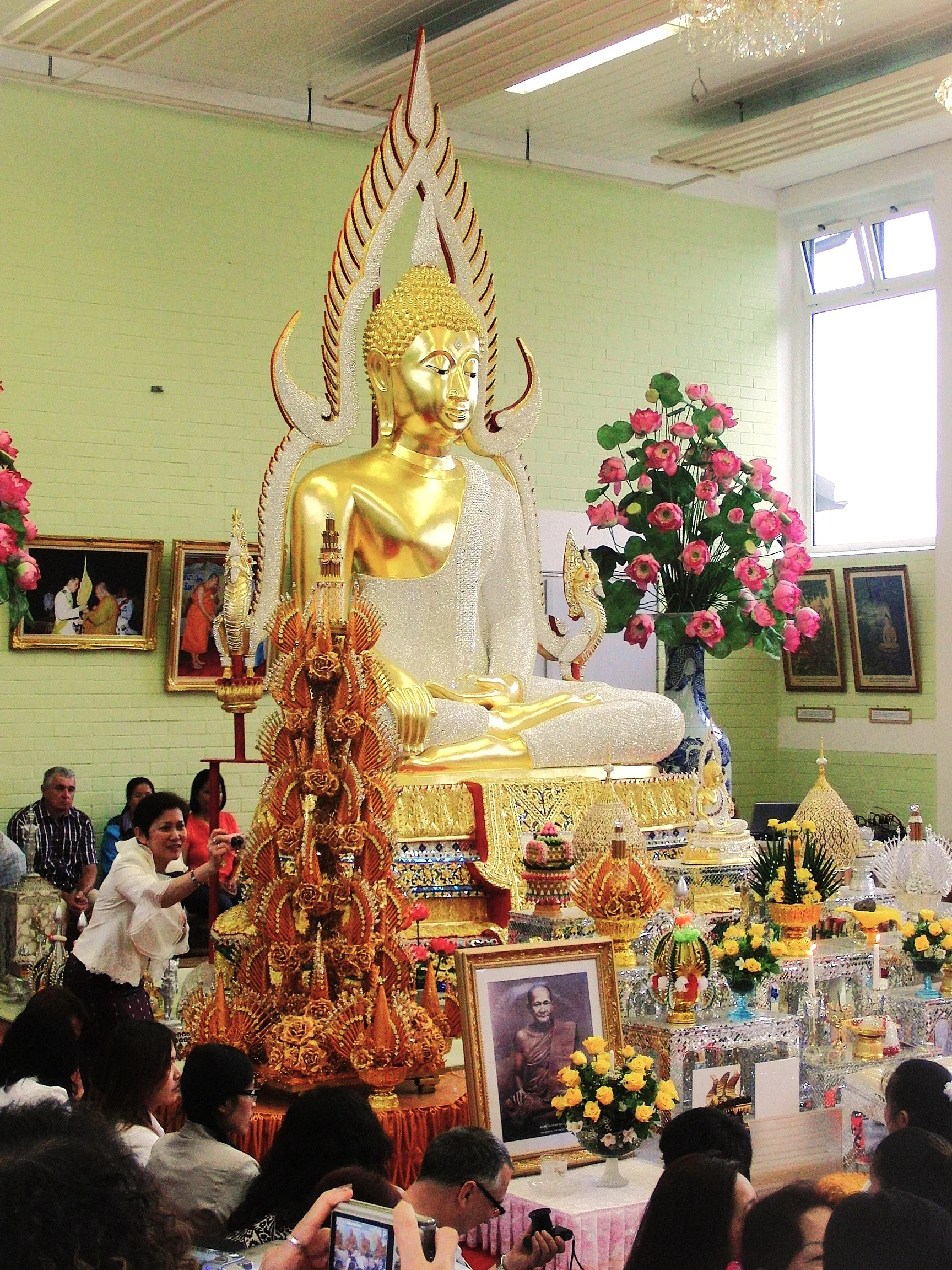 Eine große prächtig goldene Buddha-Statue umgeben von Blumenschmuck und meditierende Menschen in einer großen Halle.