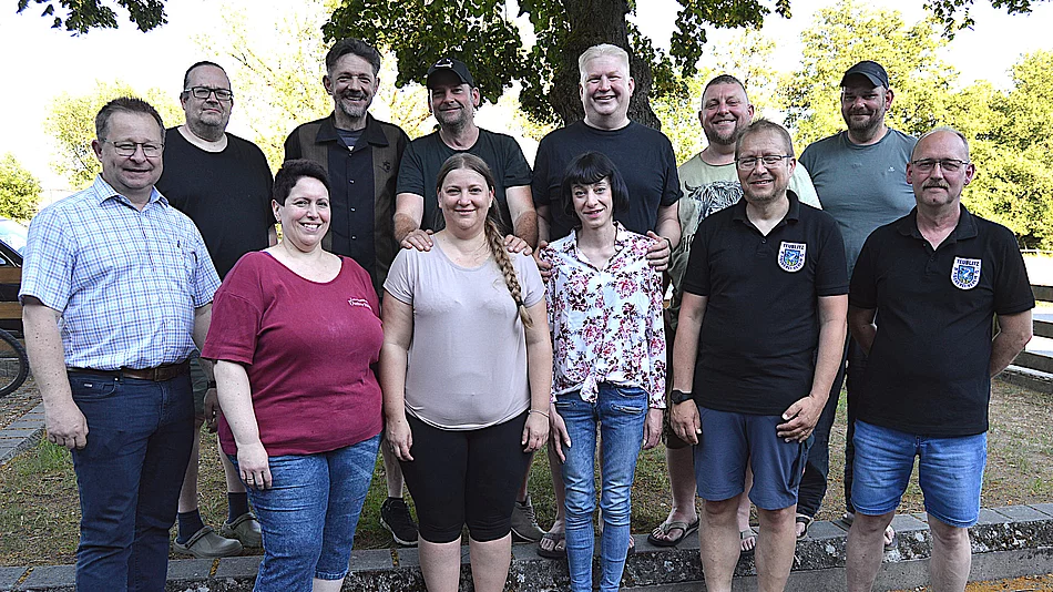 Elf Personen stehen für ein Gruppenfoto im Freien vor Bäumen und einem Zaun, wobei einige Freizeitkleidung und zwei schwarze Poloshirts mit Teublitzer Wappen tragen und gemeinsam in die Kamera blicken.