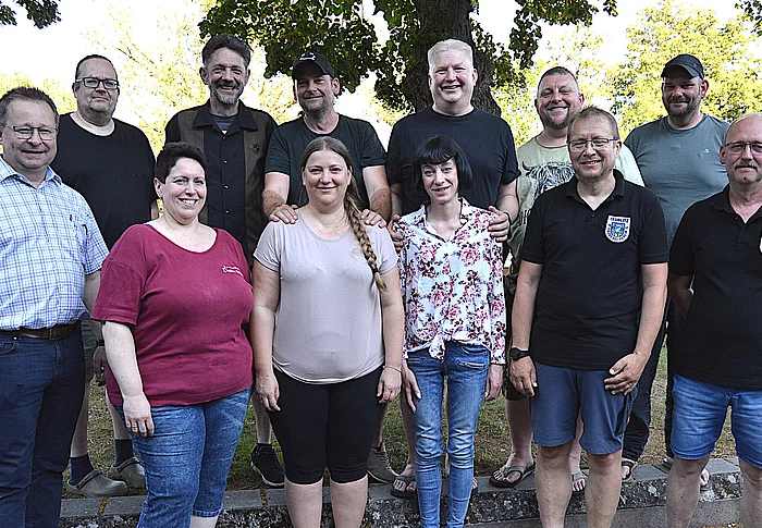 Elf Personen stehen für ein Gruppenfoto im Freien vor Bäumen und einem Zaun, wobei einige Freizeitkleidung und zwei schwarze Poloshirts mit Teublitzer Wappen tragen und gemeinsam in die Kamera blicken.