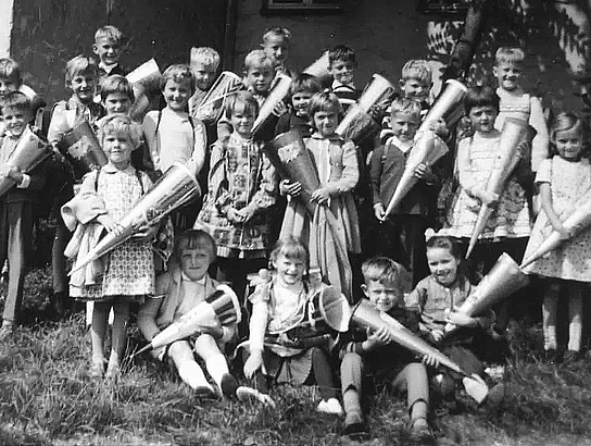 Historische Schwarz-Weiß-Aufnahme der Erstklässler in Münchshofen im Schuljahr 1963/64. Die Kinder stehen mit ihren Schultüten in mehreren Reihen vor dem Schulgebäude.