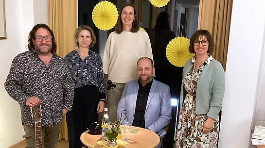 Fünf Personen stehen und sitzen in einem gemütlichen Veranstaltungsraum um einen kleinen Tisch mit Lampe, Wasserflasche und Dekoration; neben ihnen lehnt eine Gitarre, während im Hintergrund gelbe Papierdekorationen an einem Fenster hängen, was auf eine musikalische Lesung oder kulturelle Veranstaltung hinweist.