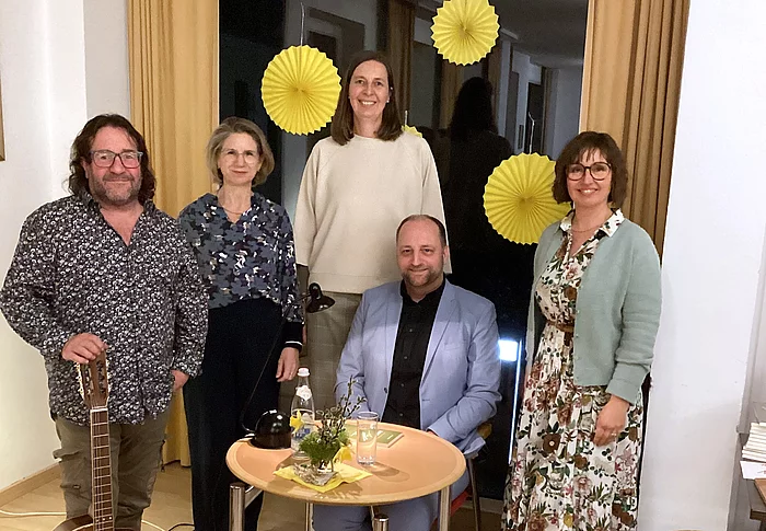 Fünf Personen stehen und sitzen in einem gemütlichen Veranstaltungsraum um einen kleinen Tisch mit Lampe, Wasserflasche und Dekoration; neben ihnen lehnt eine Gitarre, während im Hintergrund gelbe Papierdekorationen an einem Fenster hängen, was auf eine musikalische Lesung oder kulturelle Veranstaltung hinweist.