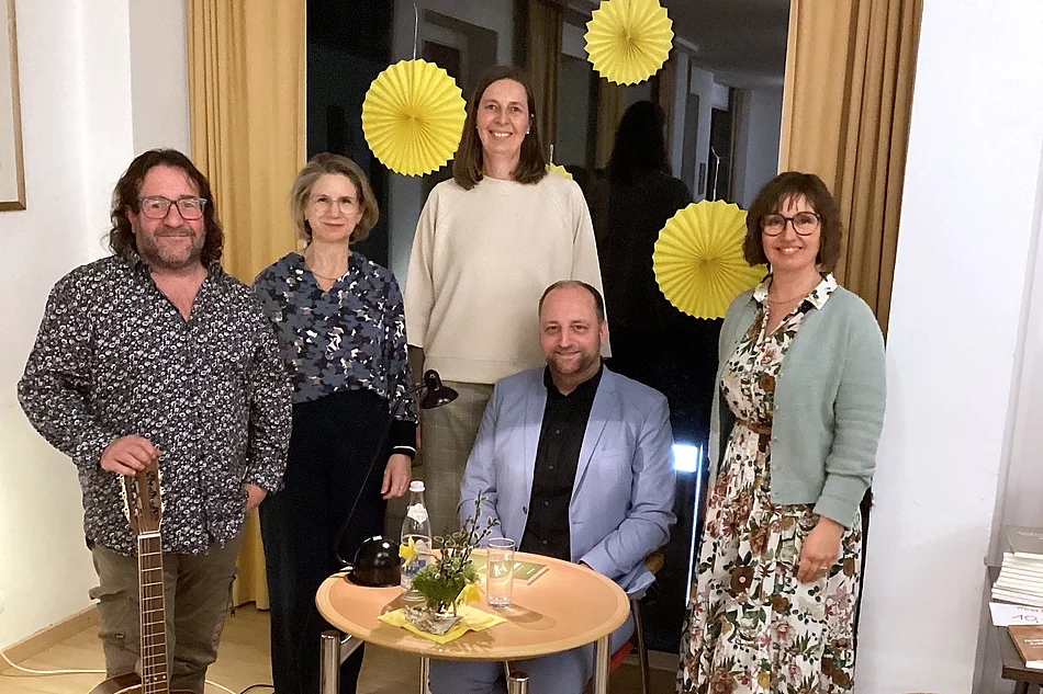 Fünf Personen stehen und sitzen in einem gemütlichen Veranstaltungsraum um einen kleinen Tisch mit Lampe, Wasserflasche und Dekoration; neben ihnen lehnt eine Gitarre, während im Hintergrund gelbe Papierdekorationen an einem Fenster hängen, was auf eine musikalische Lesung oder kulturelle Veranstaltung hinweist.