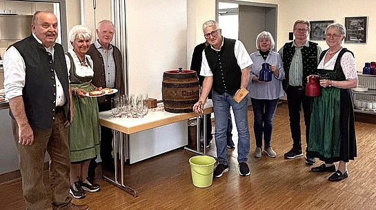 Sieben Seniorinnen und Senioren stehen beim Oktoberfest im Mehrgenerationenhaus rund um ein kleines Holzfass, einige tragen Tracht und halten Kannen oder bayerische Schmankerl in den Händen.
