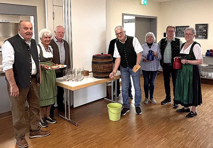 Sieben Seniorinnen und Senioren stehen beim Oktoberfest im Mehrgenerationenhaus rund um ein kleines Holzfass, einige tragen Tracht und halten Kannen oder bayerische Schmankerl in den Händen.