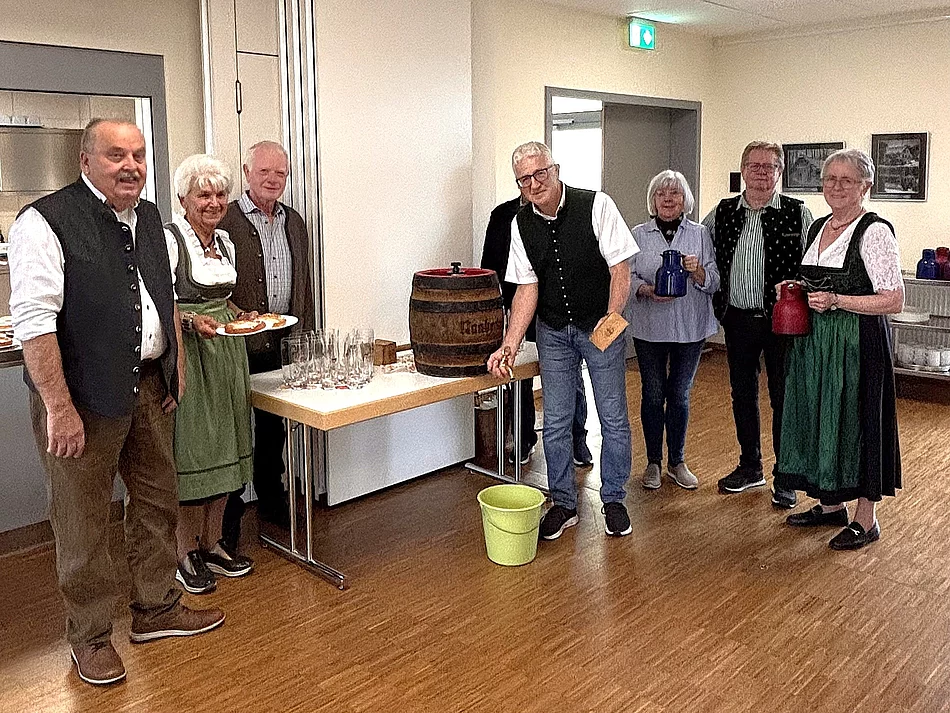 Sieben Seniorinnen und Senioren stehen beim Oktoberfest im Mehrgenerationenhaus rund um ein kleines Holzfass, einige tragen Tracht und halten Kannen oder bayerische Schmankerl in den Händen.