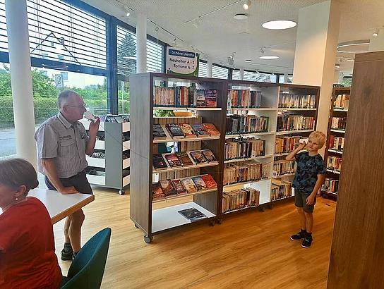 In der Stadtbücherei stehen sich ein älterer Mann und ein Junge mit etwas Abstand gegenüber. Beide halten jeweils einen Becher an ihr Ohr; zwischen ihnen verläuft eine gespannte Schnur.
