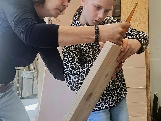 Eine erwachsene Frau und ein Mädchen arbeiten gemeinsam an einem länglichen Holzbrett. Die Frau zeigt dem Mädchen die Handhabung eines Werkzeugs.