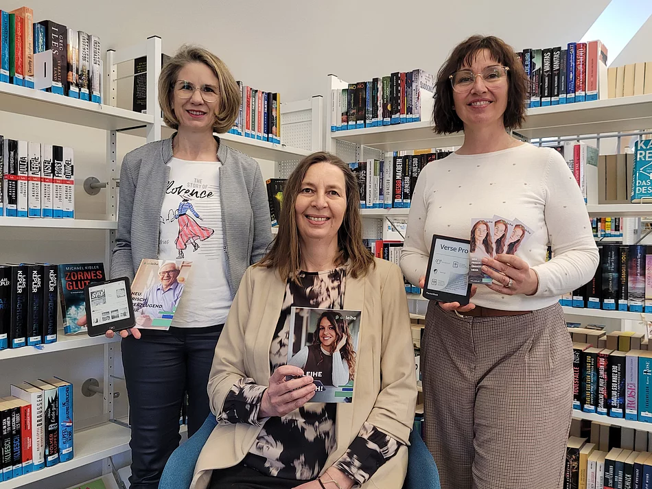 Drei Frauen stehen und sitzen zwischen Bücherregalen in einer Bibliothek und halten verschiedene Medien wie ein E-Book-Lesegerät, Flyer und Informationskarten in den Händen.