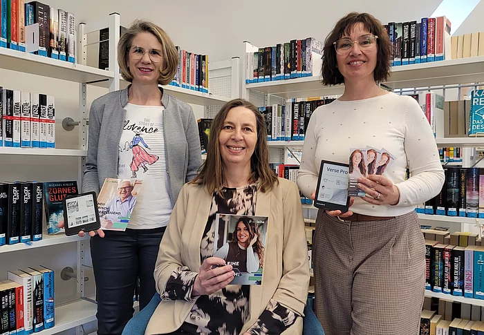 Drei Frauen stehen und sitzen zwischen Bücherregalen in einer Bibliothek und halten verschiedene Medien wie ein E-Book-Lesegerät, Flyer und Informationskarten in den Händen.