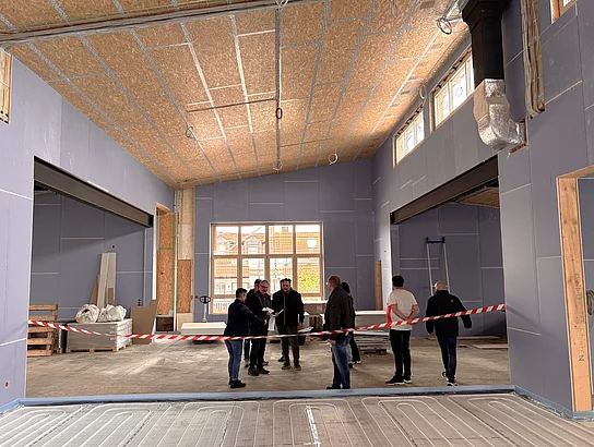 Mehrere Menschen stehen in einer großen Baustellenhalle mit hohen Decken und schauen sich den Innenraum eines im Bau befindlichen Gebäudes an.