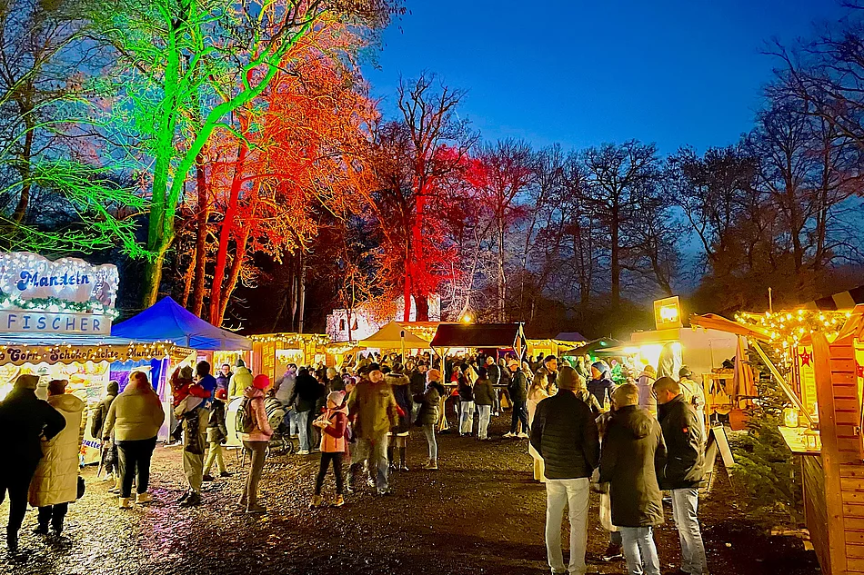 Das Bild zeigt einen lebhaften Weihnachtsmarkt am Abend, mit bunt beleuchteten Bäumen, festlich geschmückten Buden, vielen Besuchern und einer warmen, fröhlichen Atmosphäre.