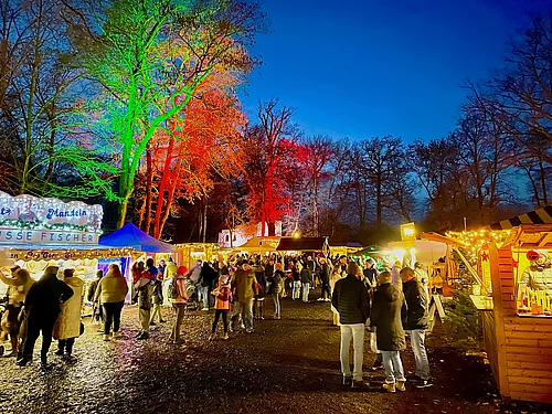 Das Bild zeigt einen lebhaften Weihnachtsmarkt am Abend, mit bunt beleuchteten Bäumen, festlich geschmückten Buden, vielen Besuchern und einer warmen, fröhlichen Atmosphäre.