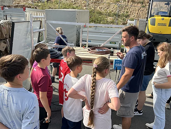 Ein Betreuer erklärt einer Gruppe von Kindern und Jugendlichen im Freien ein technisches Gerät. Im Hintergrund stehen ein Anhänger, Kabel und weitere Arbeitsgeräte.