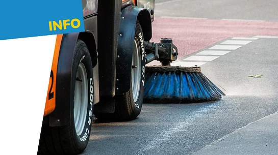 Nahaufnahme einer Kehrmaschine bei der Straßenreinigung. Zu sehen sind die Räder des Fahrzeugs sowie eine große rotierende Bürste, die über die Fahrbahn fegt. Links oben befindet sich auf blauem Hintergrund der gelbe Schriftzug „Info“.
