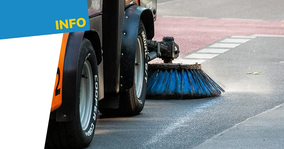 Nahaufnahme einer Kehrmaschine bei der Straßenreinigung. Zu sehen sind die Räder des Fahrzeugs sowie eine große rotierende Bürste, die über die Fahrbahn fegt. Links oben befindet sich auf blauem Hintergrund der gelbe Schriftzug „Info“.