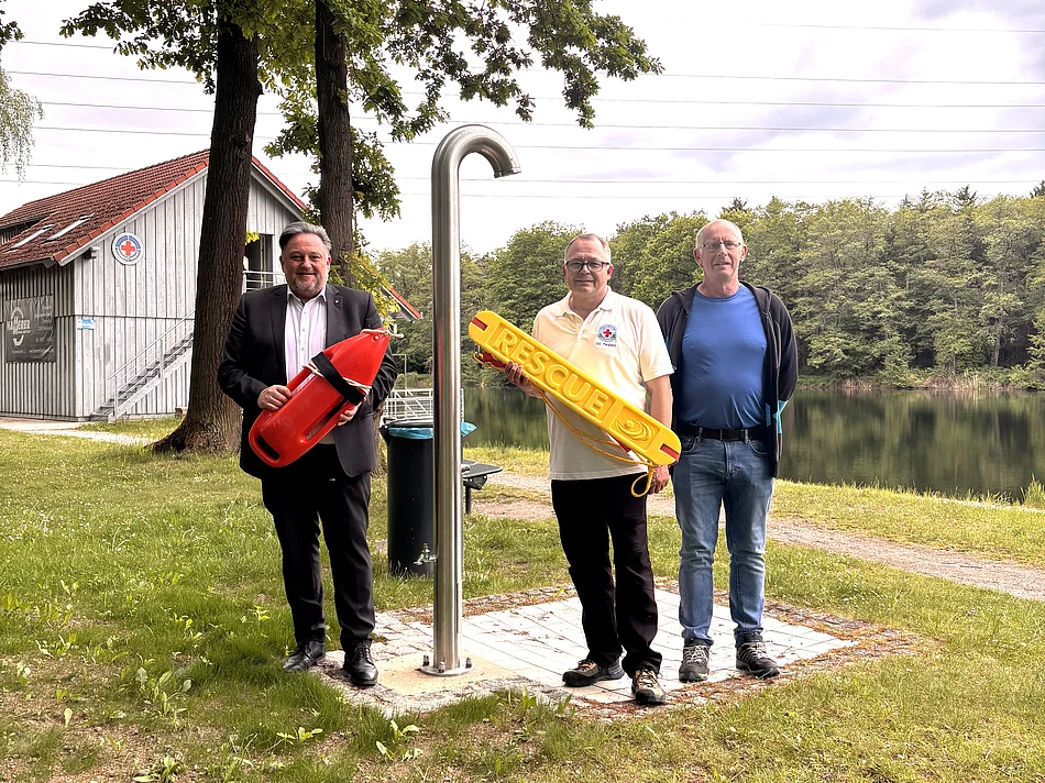 Drei Männer stehen auf einer Grünfläche am Ufer eines Sees neben einer Edelstahldusche im Freien. Einer der Männer hält eine rote Rettungsboje, ein anderer einen gelben Rettungsring mit der Aufschrift „RESCUE“. Im Hintergrund sind der ruhige See, Bäume und ein Gebäude der Wasserwacht zu sehen. Die Männer blicken in die Kamera, der Himmel ist leicht bewölkt. Die Aufnahme vermittelt den Eindruck eines Termins zum Start der Badesaison oder zur Vorstellung von Sicherheitsausstattung am Badesee.