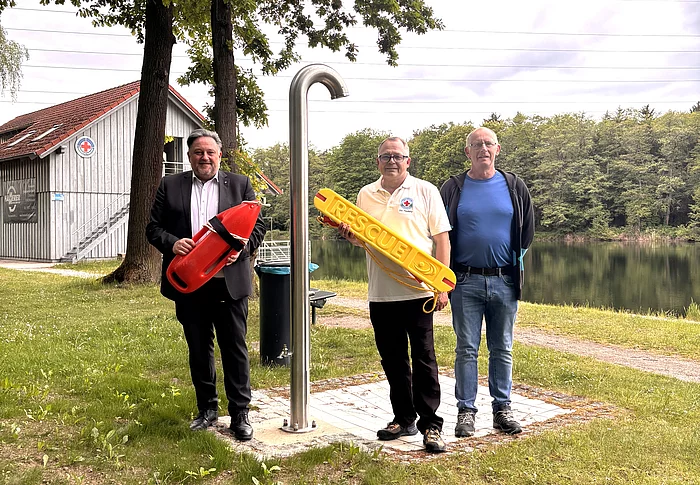 Drei Männer stehen auf einer Grünfläche am Ufer eines Sees neben einer Edelstahldusche im Freien. Einer der Männer hält eine rote Rettungsboje, ein anderer einen gelben Rettungsring mit der Aufschrift „RESCUE“. Im Hintergrund sind der ruhige See, Bäume und ein Gebäude der Wasserwacht zu sehen. Die Männer blicken in die Kamera, der Himmel ist leicht bewölkt. Die Aufnahme vermittelt den Eindruck eines Termins zum Start der Badesaison oder zur Vorstellung von Sicherheitsausstattung am Badesee.