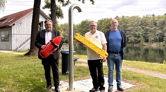 Drei Männer stehen auf einer Grünfläche am Ufer eines Sees neben einer Edelstahldusche im Freien. Einer der Männer hält eine rote Rettungsboje, ein anderer einen gelben Rettungsring mit der Aufschrift „RESCUE“. Im Hintergrund sind der ruhige See, Bäume und ein Gebäude der Wasserwacht zu sehen. Die Männer blicken in die Kamera, der Himmel ist leicht bewölkt. Die Aufnahme vermittelt den Eindruck eines Termins zum Start der Badesaison oder zur Vorstellung von Sicherheitsausstattung am Badesee.