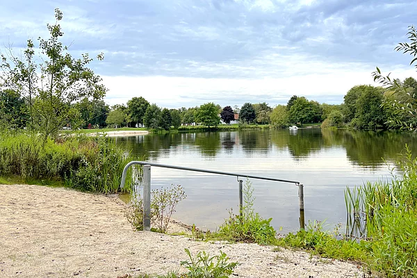 Ufereinstieg an einem Badesee mit Sandbereich, grünem Uferbewuchs und Handlauf.