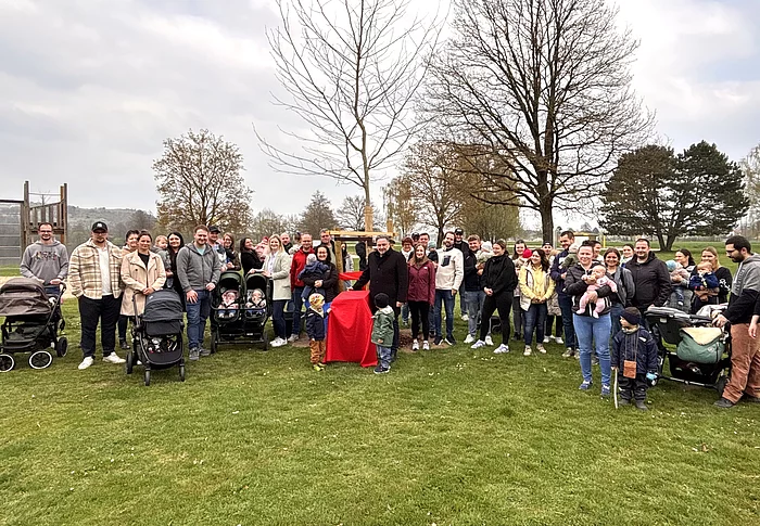 Eine große Gruppe von Familien mit Kindern und Kinderwagen versammelt sich auf einer Wiese rund um einen frisch gepflanzten Baum.