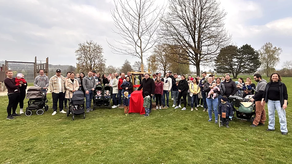 Eine große Gruppe von Familien mit Kindern und Kinderwagen versammelt sich auf einer Wiese rund um einen frisch gepflanzten Baum.