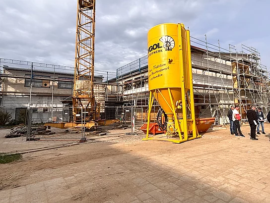 Auf einer Baustelle mit Baukran und großem gelben Baustoffsilo stehen mehrere Personen und betrachten das im Bau befindliche Gebäude.