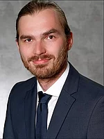 Ein Mann mit zurückgebundenem Haar und Bart trägt einen Anzug mit Krawatte und blickt mit leichtem Lächeln vor neutralem Hintergrund in die Kamera.