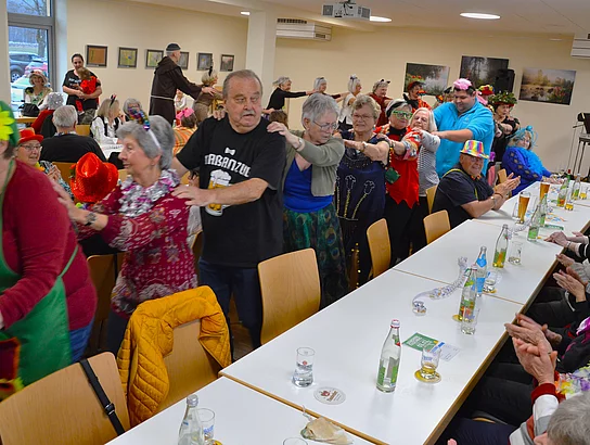 Eine lange Polonaise aus Seniorinnen und Senioren zieht lachend und Schulter an Schulter durch den Raum zwischen den Tischen.