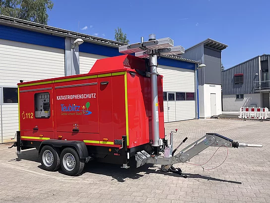 Ein mobiles rotes Notstromaggregat auf einem Anhänger steht auf dem Gelände eines Wasserwerks. Die Seitenklappe ist geöffnet und gibt den Blick auf die technische Steuerungs- und Schaltanlage frei. Auf dem Fahrzeug sind reflektierende Warnmarkierungen sowie die Aufschrift „Katastrophenschutz Teublitz“ zu sehen.