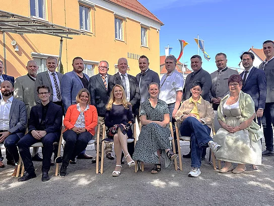 Eine größere Gruppe von Frauen und Männern sitzt und steht gemeinsam vor einem Gebäude und posiert bei sonnigem Wetter für ein Gruppenfoto.